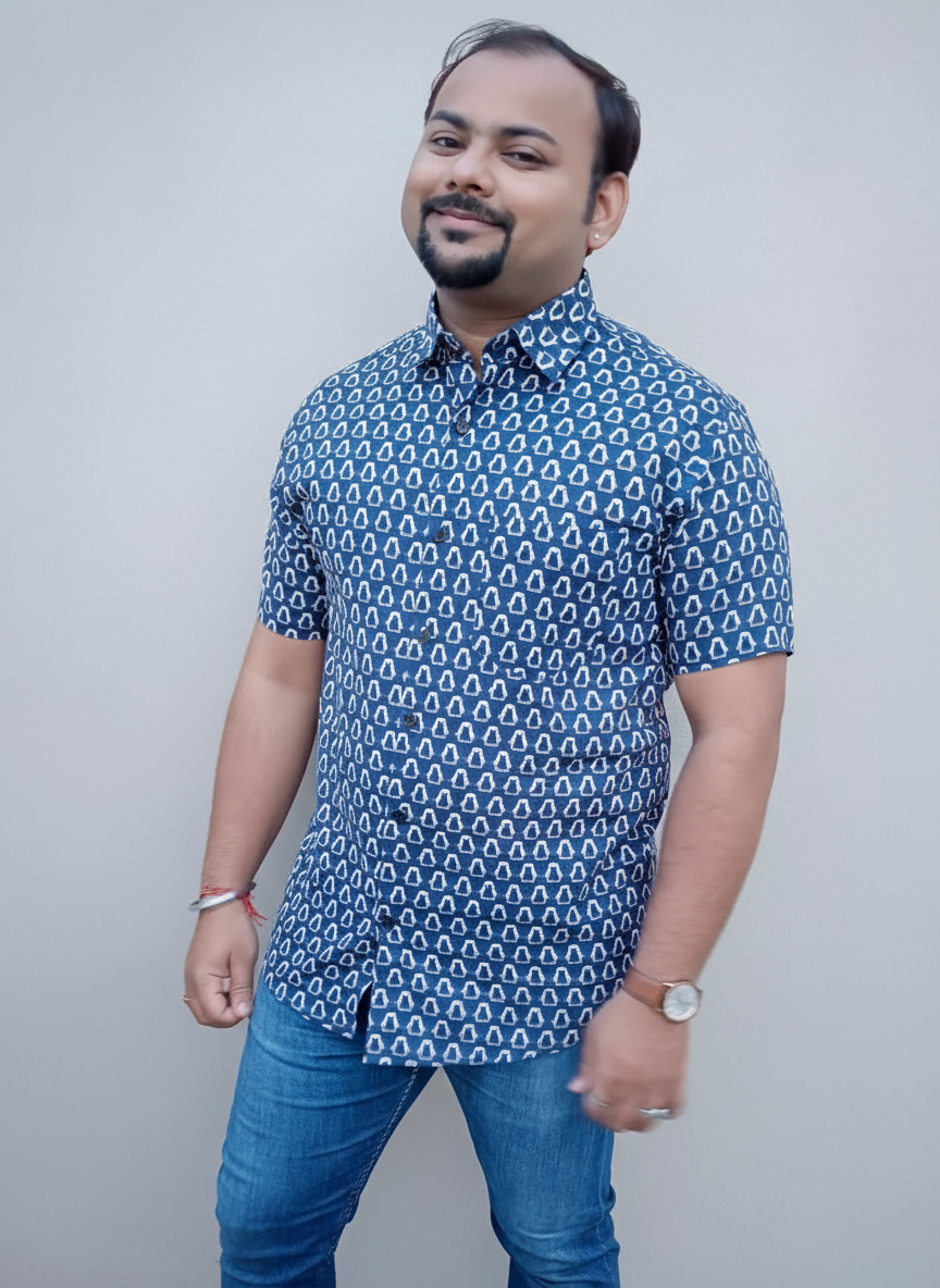 Man wearing a blue patterned shirt and jeans standing against a plain wall.
