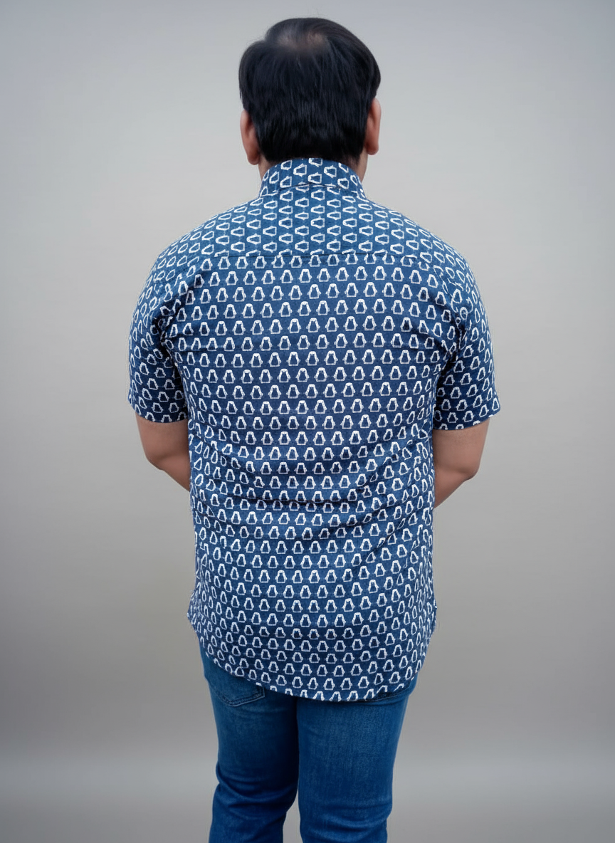 Person wearing a blue patterned shirt and jeans standing against a plain wall.