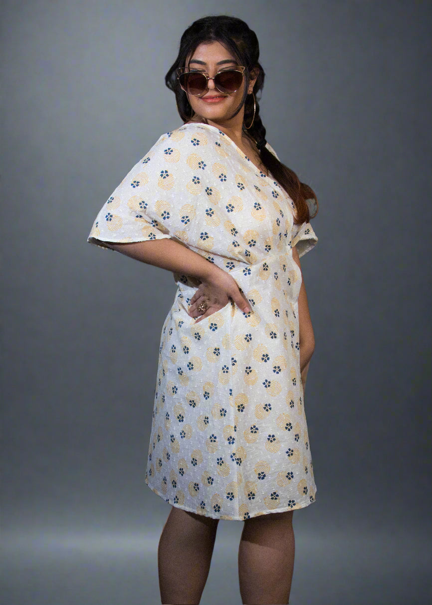 Woman wearing a white floral dress standing against a black background with a plant to her left.