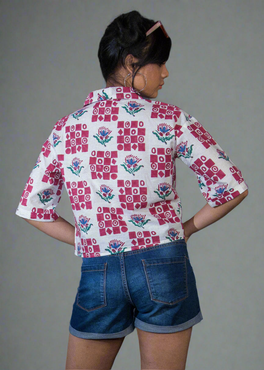 Person wearing a patterned shirt and denim shorts standing against a dark backdrop with a plant on the left.