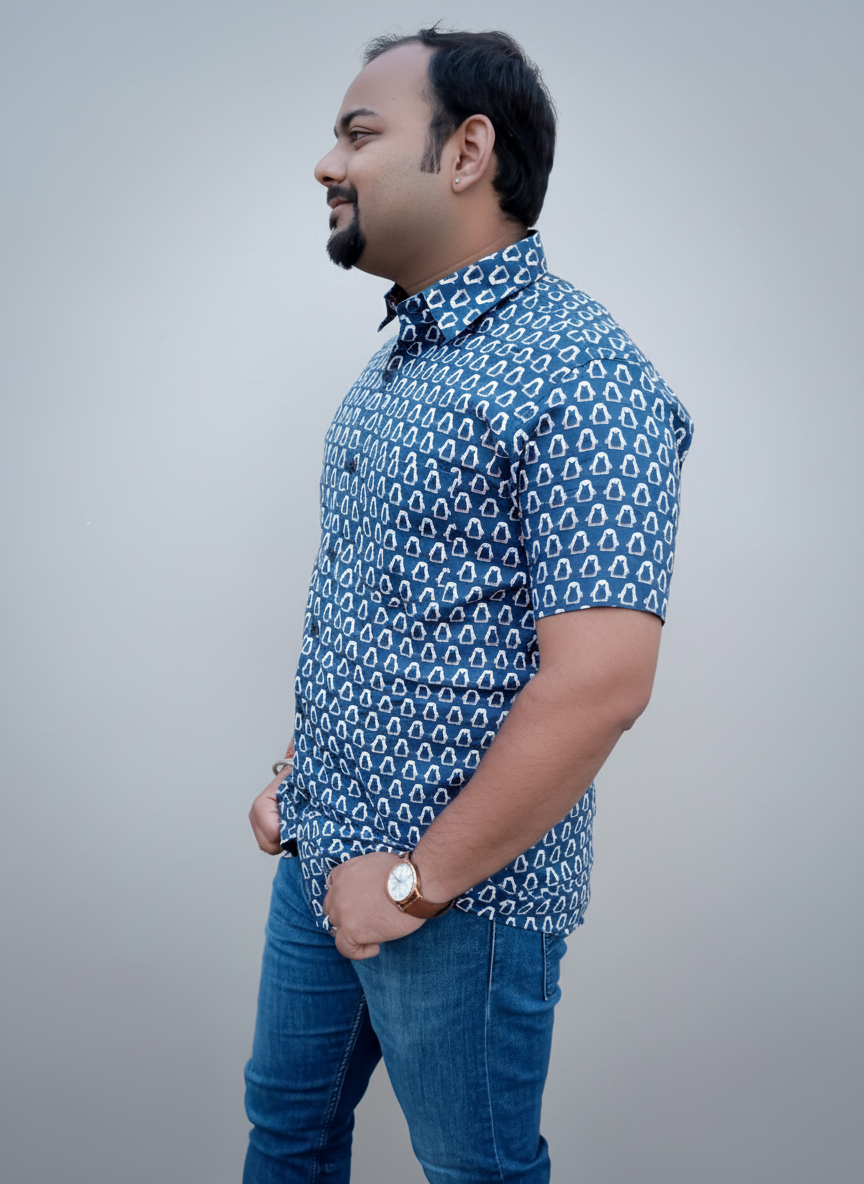 Man wearing a blue patterned shirt and jeans standing against a plain wall.