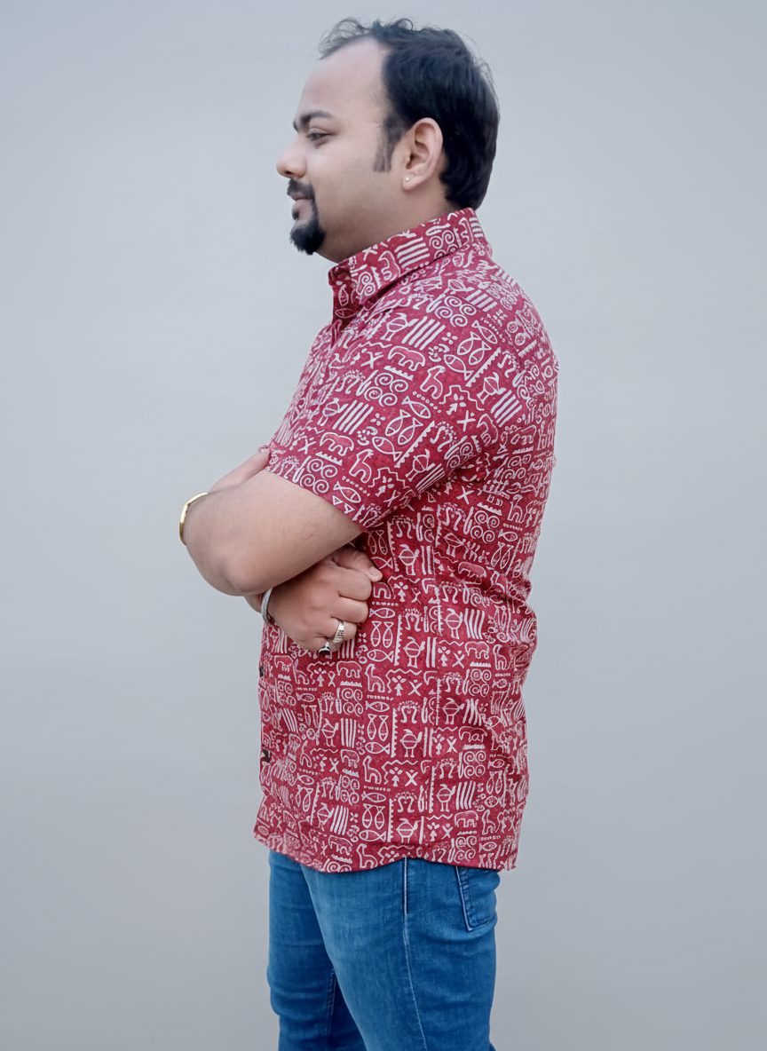 Man wearing a red patterned shirt and blue jeans standing against a plain wall.