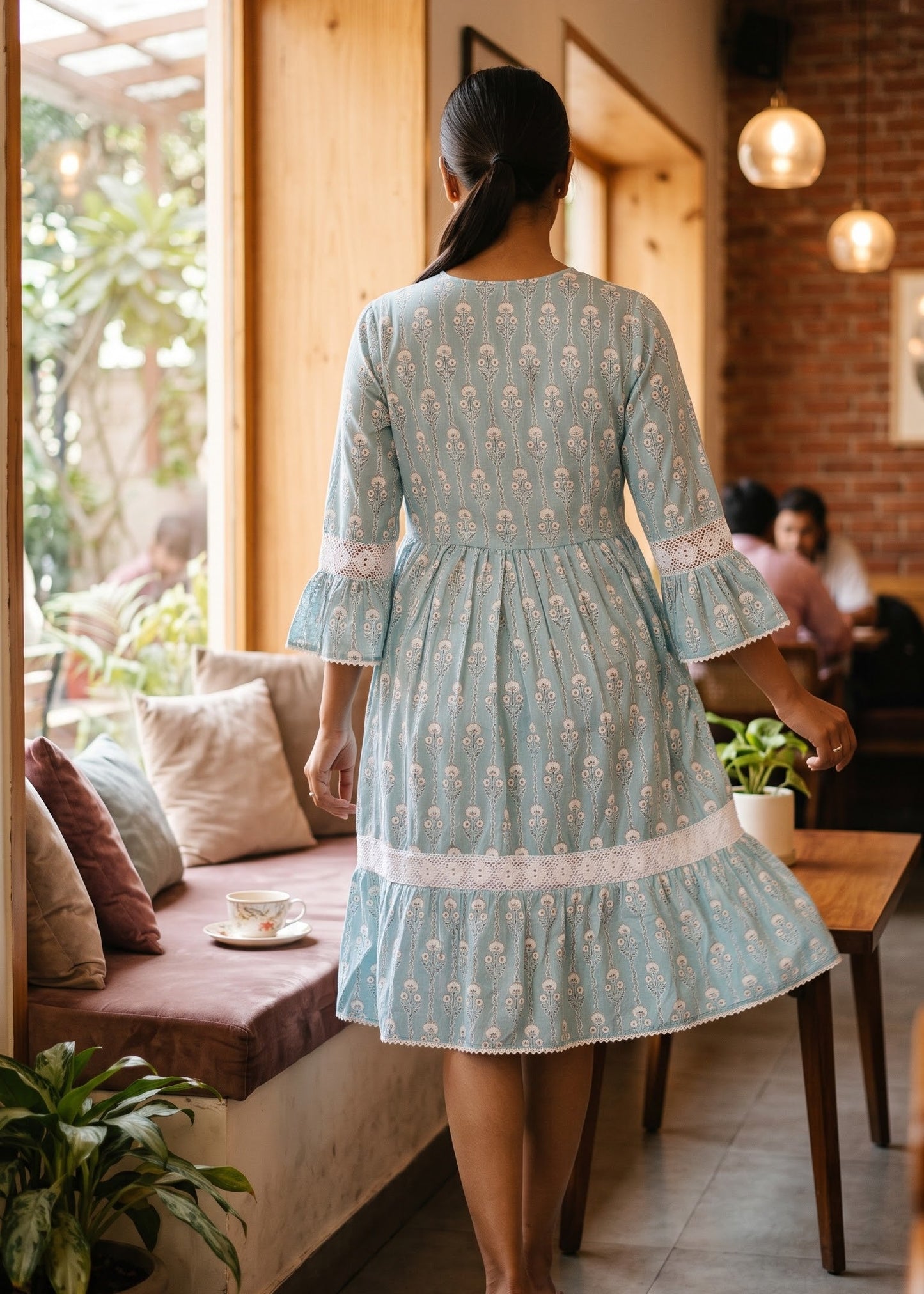 A woman viewed from behind wearing a light blue and white printed cotton dress with bell sleeves, standing in a cozy cafe interior with wooden windows, cushions, and pendant lights.