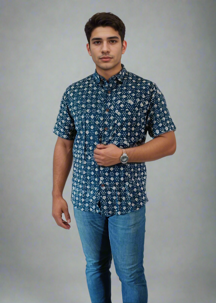 A man standing against a grey background, wearing a blue indigo daabu print cotton shirt with half sleeves and blue jeans.