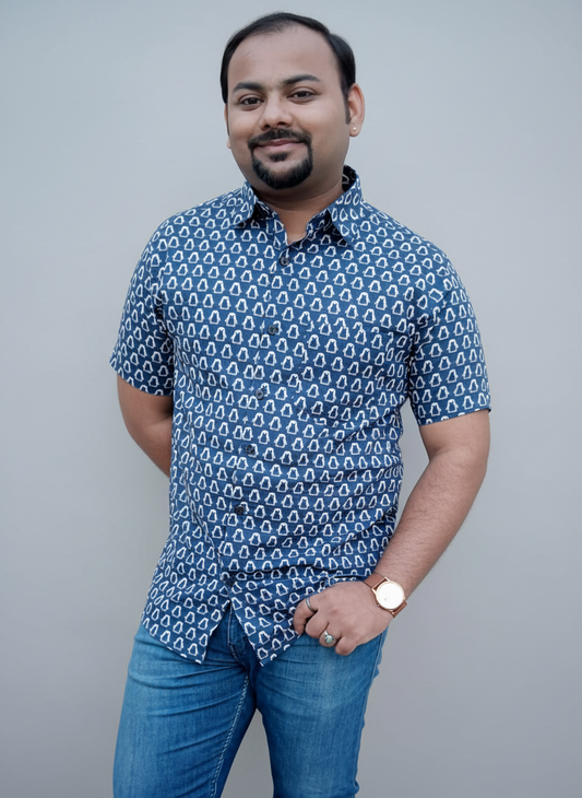 Man wearing a blue patterned shirt and jeans standing against a plain wall.
