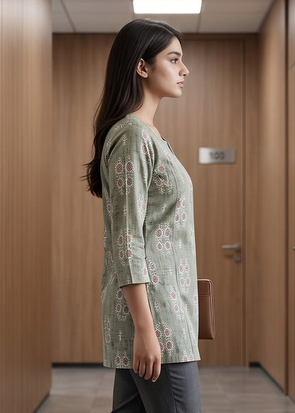 A woman walks down a modern office hallway wearing a light sage green patterned tunic top, dark gray trousers, and brown loafers.