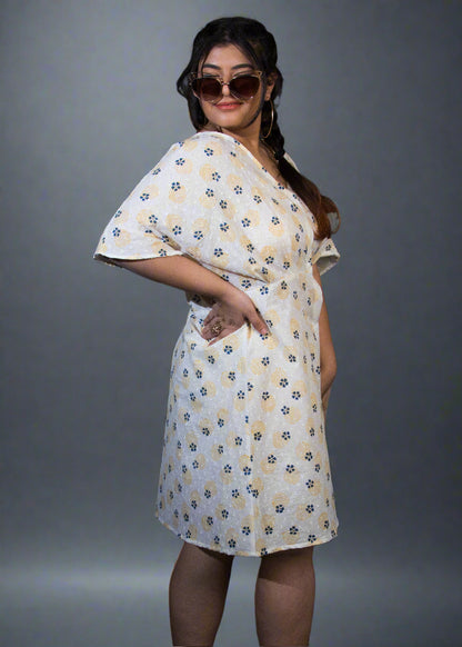 Woman wearing a white floral dress standing against a black background with a plant to her left.
