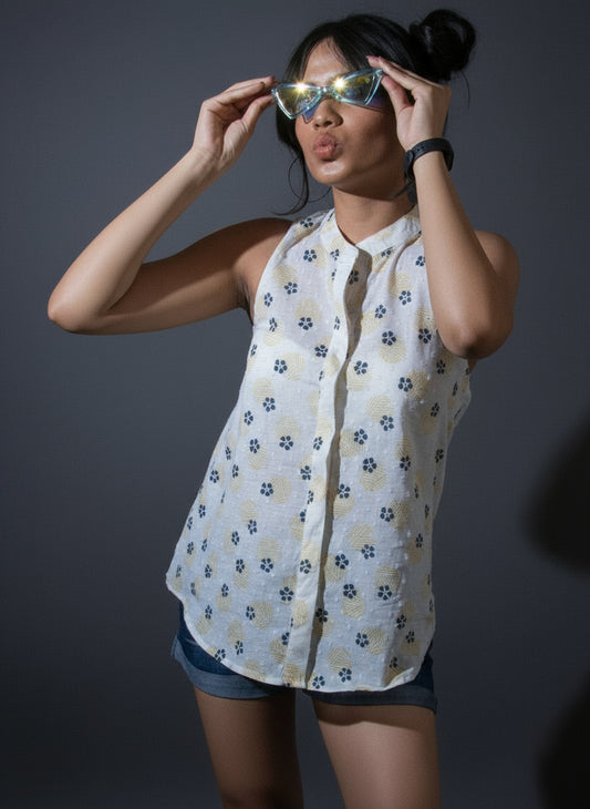 Person wearing a patterned shirt and shorts standing against a dark curtain with a plant on the left.