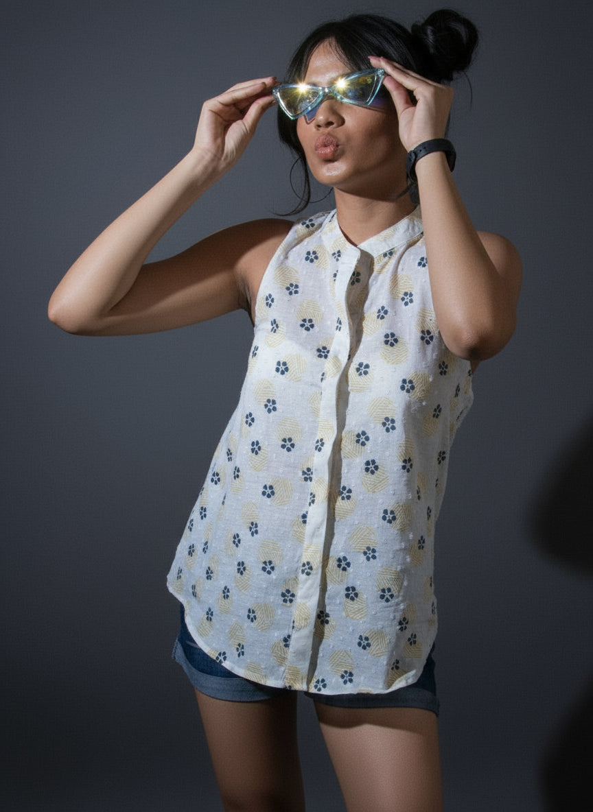 Person wearing a patterned shirt and shorts standing against a dark curtain with a plant on the left.