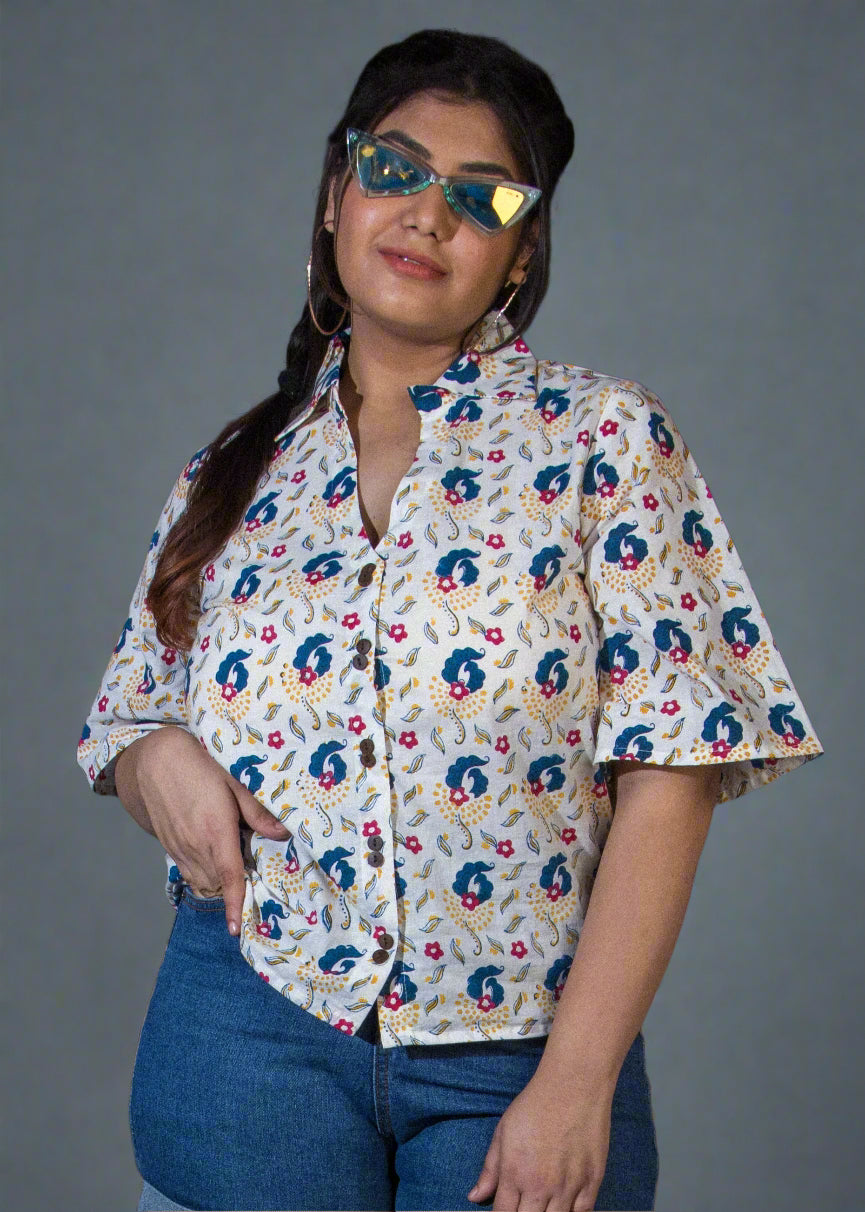 Person wearing a hand block floral shirt and denim shorts standing against a black backdrop with a plant to the side.