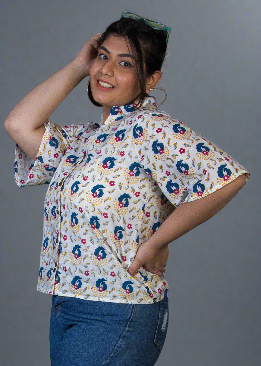 Person wearing a floral shirt and denim shorts standing against a black backdrop with a plant on the left.