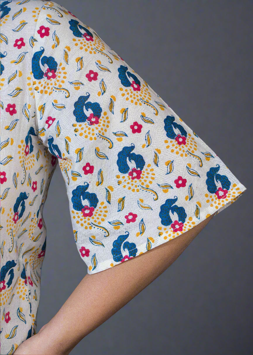 Person wearing a hand block floral paisley shirt and denim shorts standing against a dark backdrop with a plant.
