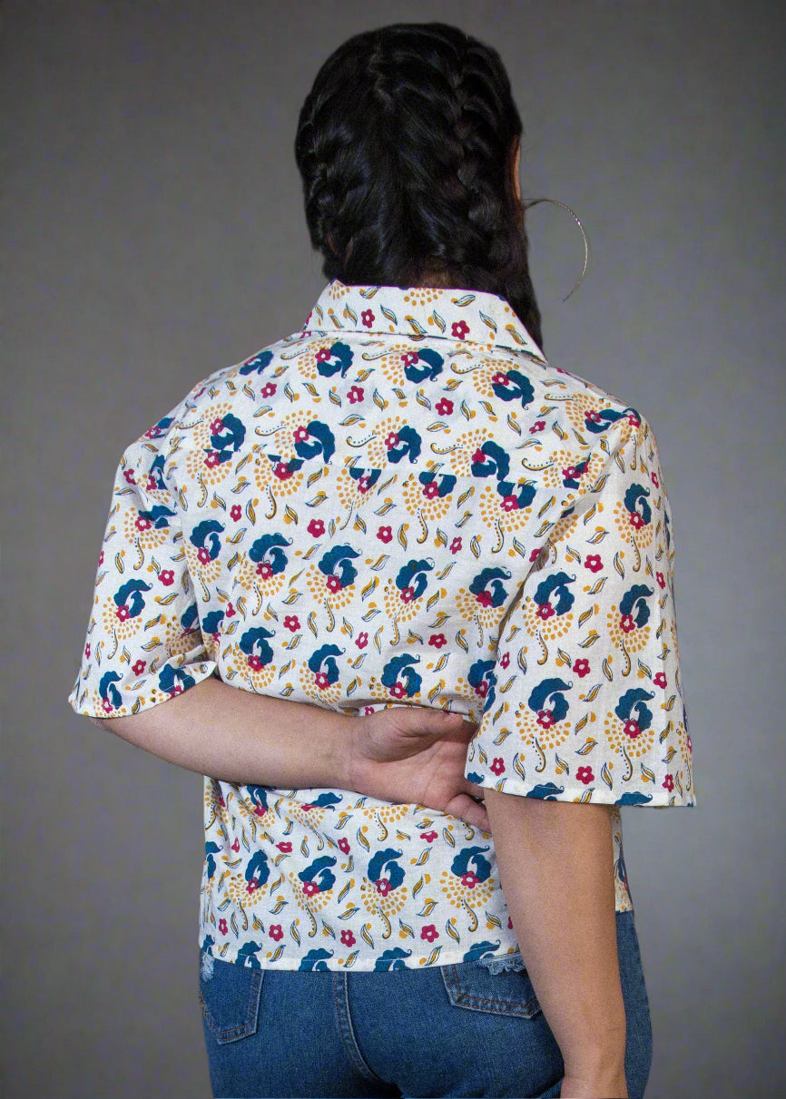 Person wearing a patterned shirt and denim shorts standing against a dark backdrop with a plant.