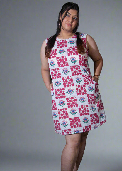 Woman wearing a patterned dress standing against a dark background with a plant to her left.