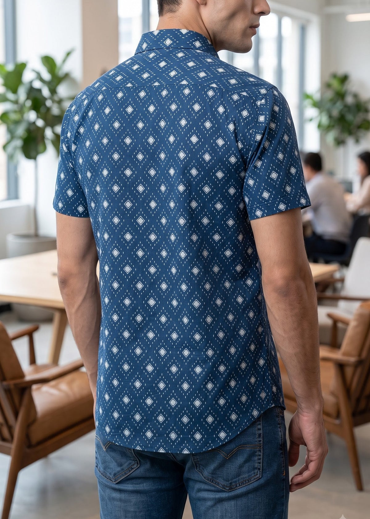 A man wearing a blue short-sleeve button-up shirt with a white geometric diamond pattern, shown from behind in a bright, modern office setting.