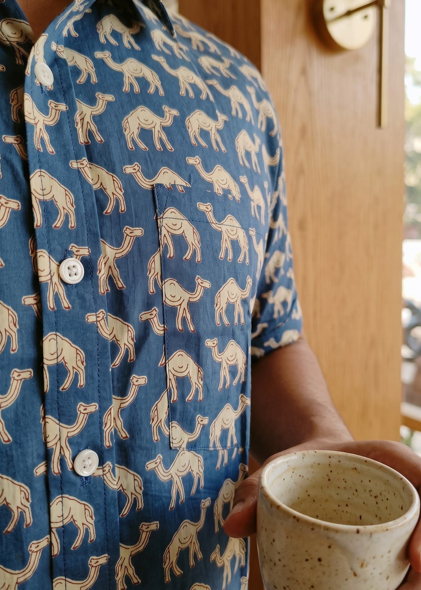 A person wearing a blue short-sleeve button-up shirt with a beige camel print, holding a speckled ceramic mug in a warm indoor setting.