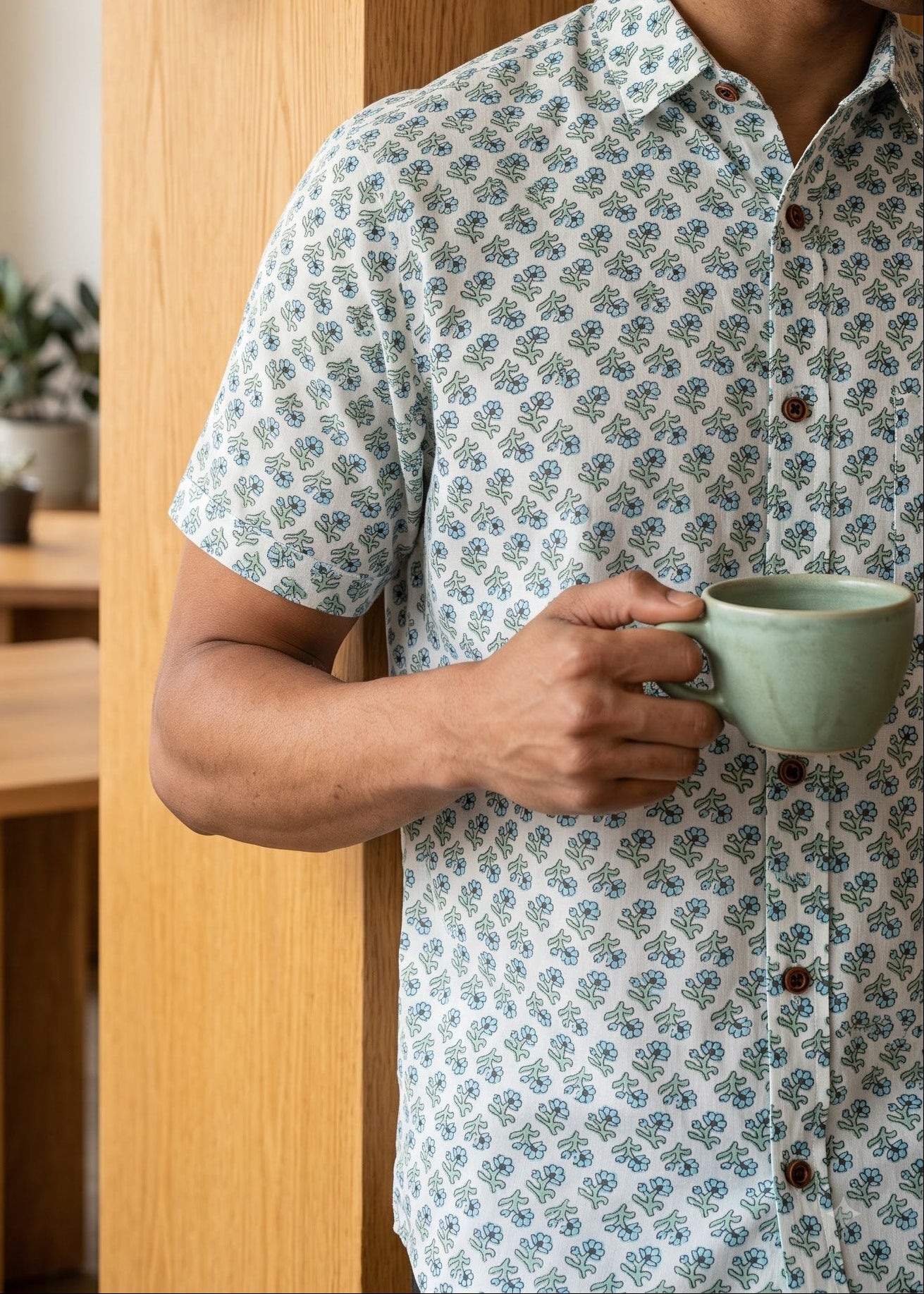 A man in a light blue and white patterned short-sleeve button-up shirt holds a sage green ceramic mug next to a wooden wall sconce in a modern, sunlit room.