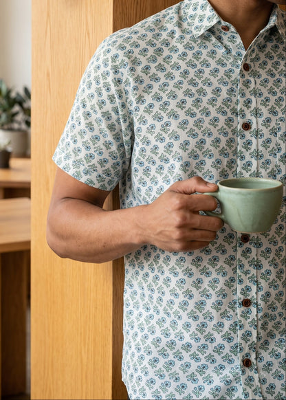 A man in a light blue and white patterned short-sleeve button-up shirt holds a sage green ceramic mug next to a wooden wall sconce in a modern, sunlit room.