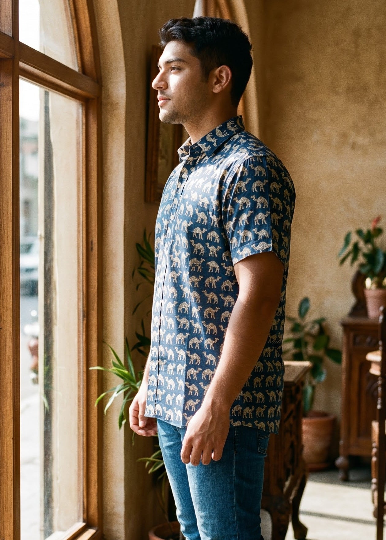A man wearing a blue and white patterned short-sleeve button-up shirt, blue jeans, and white sneakers stands in profile by a large arched window in a rustic indoor setting.