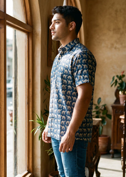 A man wearing a blue and white patterned short-sleeve button-up shirt, blue jeans, and white sneakers stands in profile by a large arched window in a rustic indoor setting.