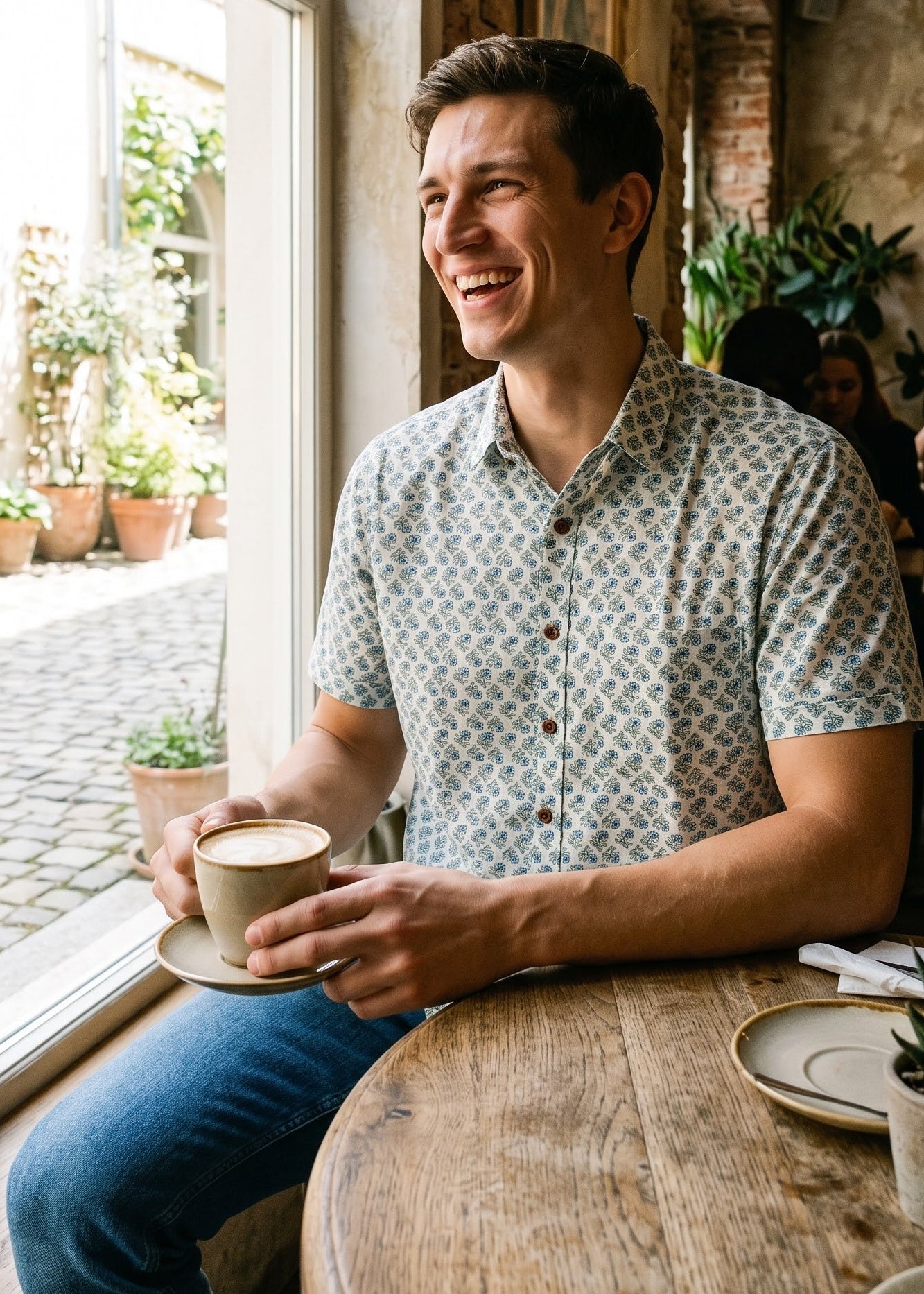 A man smiling while sitting at a wooden table in a cafe, holding a coffee cup. He is wearing a short-sleeve button-up shirt with a blue and white floral pattern and blue jeans, set against a warm, rustic interior with a large arched window.
