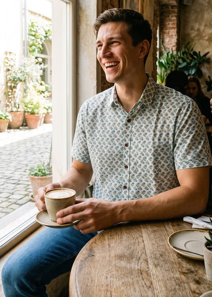 A man smiling while sitting at a wooden table in a cafe, holding a coffee cup. He is wearing a short-sleeve button-up shirt with a blue and white floral pattern and blue jeans, set against a warm, rustic interior with a large arched window.