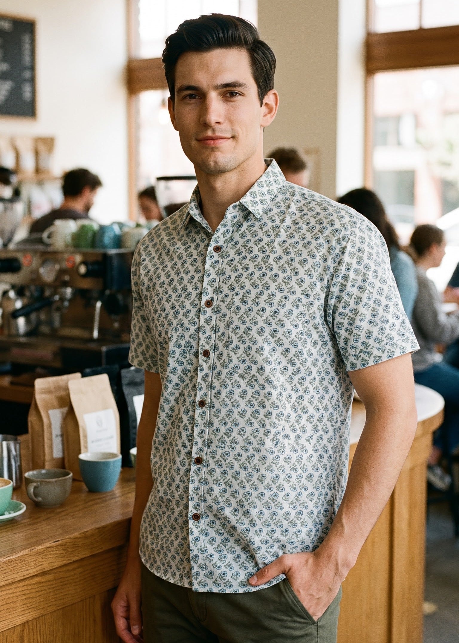 A man standing in a cafe wearing a short-sleeve button-up shirt with a light blue and white floral pattern, paired with olive green pants.