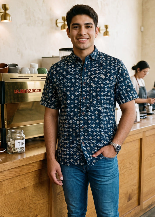 A man wearing a navy indigo blue short-sleeve button-up shirt with a white geometric pattern, blue jeans, and white sneakers stands in a coffee shop.