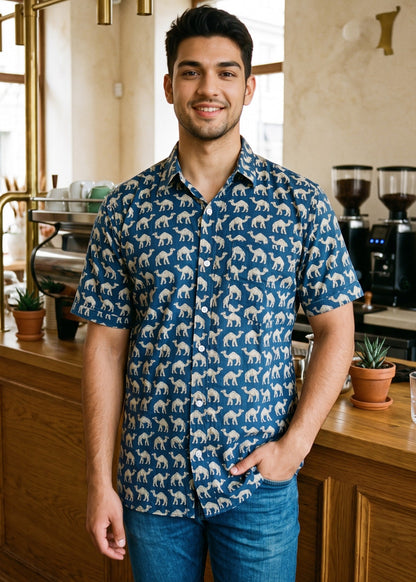 A man wearing a blue short-sleeve button-up shirt with a beige camel pattern, standing in a warm, rustic coffee shop environment.