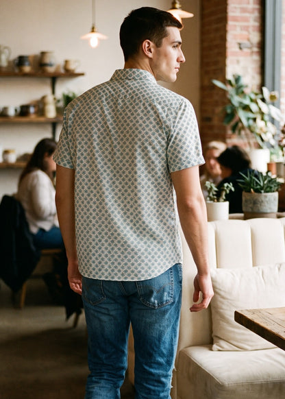 A man standing in a cafe, viewed from behind, wearing a light-colored short-sleeve button-down shirt with a small repeating floral pattern, blue jeans, and black boots.