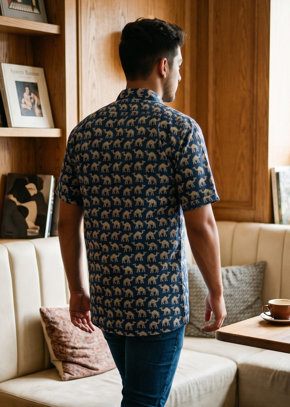A man wearing a navy blue short-sleeve button-up shirt with a beige and brown camel print, blue jeans, and white sneakers, standing in a cozy wood-paneled cafe booth.