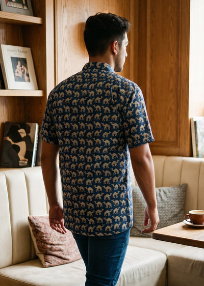 A man wearing a navy blue short-sleeve button-up shirt with a beige and brown camel print, blue jeans, and white sneakers, standing in a cozy wood-paneled cafe booth.