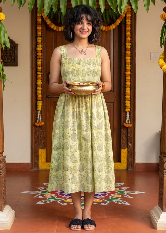 A woman stands in a decorated doorway holding a brass bowl, wearing a sleeveless light green hand block printed dress with a yellow-green leaf pattern.