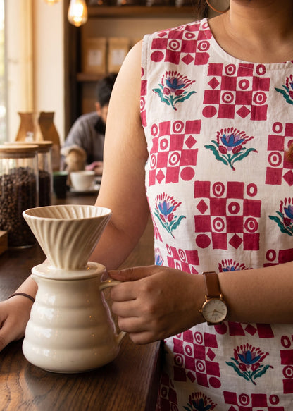 A woman in a sleeveless white dress with a red and blue geometric pattern stands at a wooden counter in a cafe, holding a white ceramic pour-over dripper and small carafe.