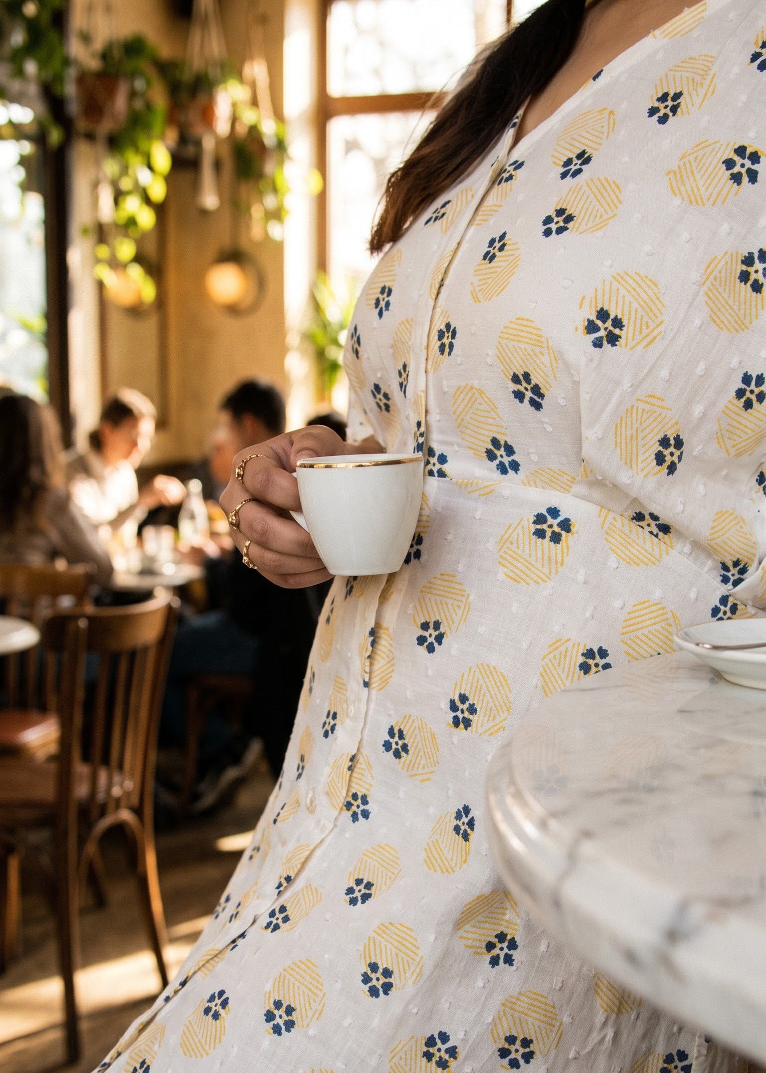 A woman in a white button down dress with a yellow and blue floral pattern holds a white ceramic cup in a sunlit cafe setting.