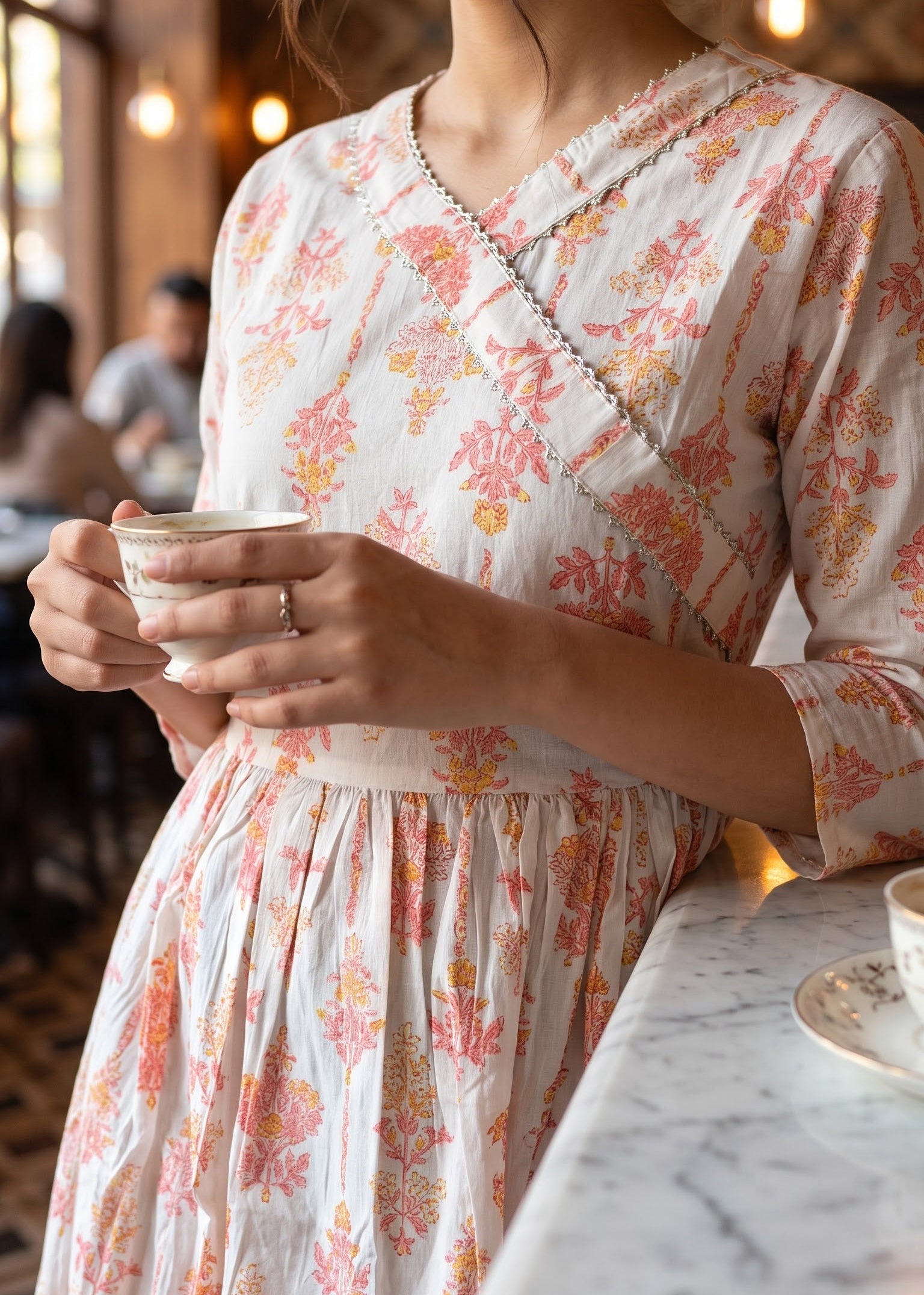 A woman in a cafe wearing a cream-colored wrap-style short kurti tunic with a pink and orange floral pattern, holding a white cup.