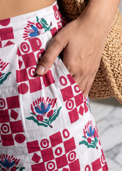 A close-up of a person wearing white pocket shorts with a red geometric checkerboard pattern and blue-and-green floral motifs.