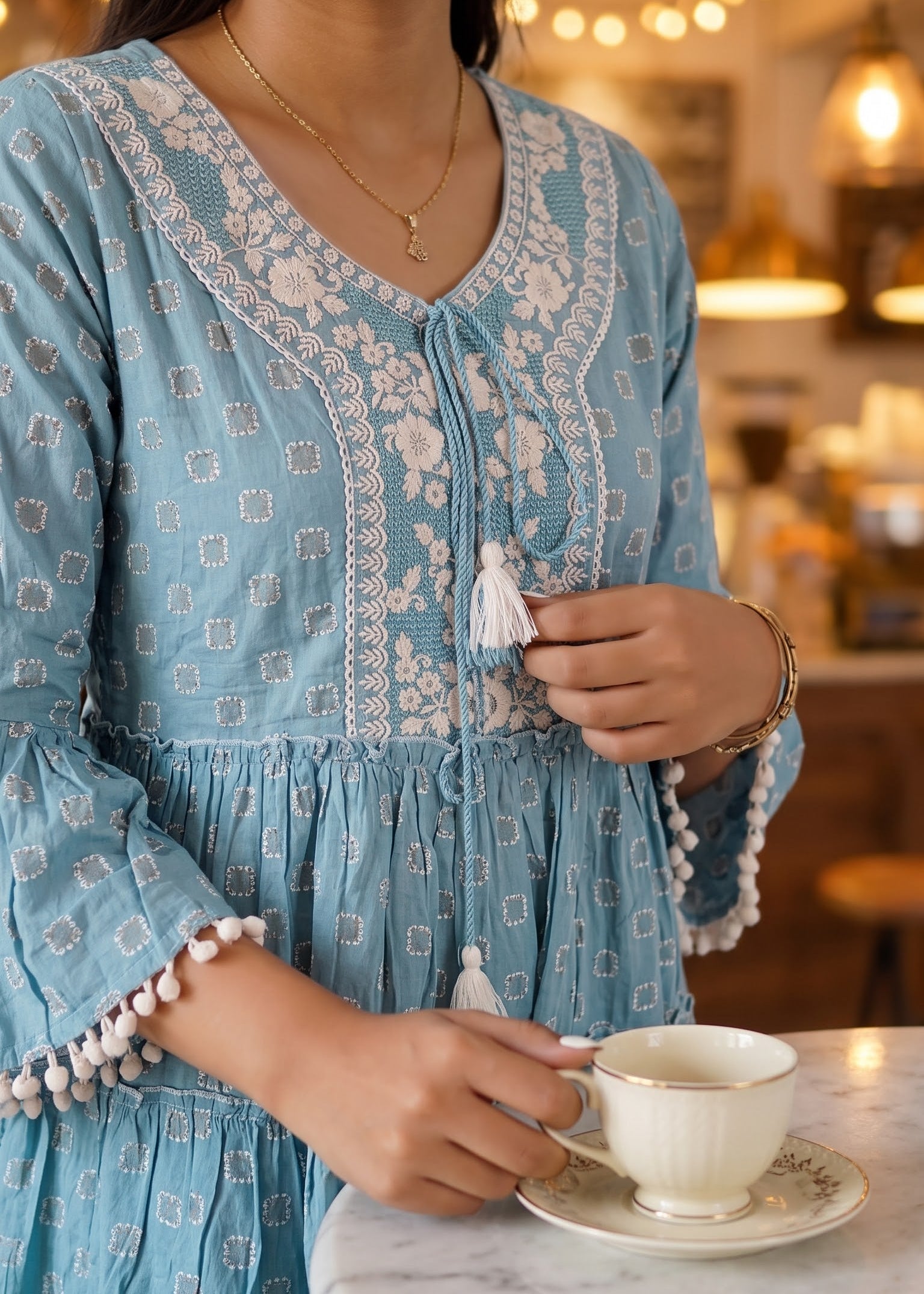 A woman wearing a light blue bohemian-style embroidered dress with white floral embroidery patterns and pom-pom trim, holding a white teacup on a saucer in a warm, cozy cafe setting.