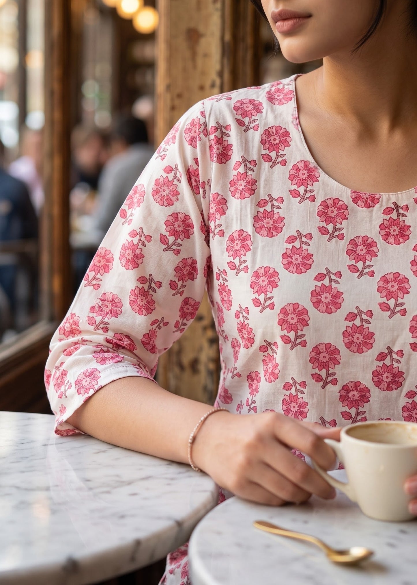 A woman wearing a white short kurta with a pink floral pattern sits at a cafe table, holding a white mug.