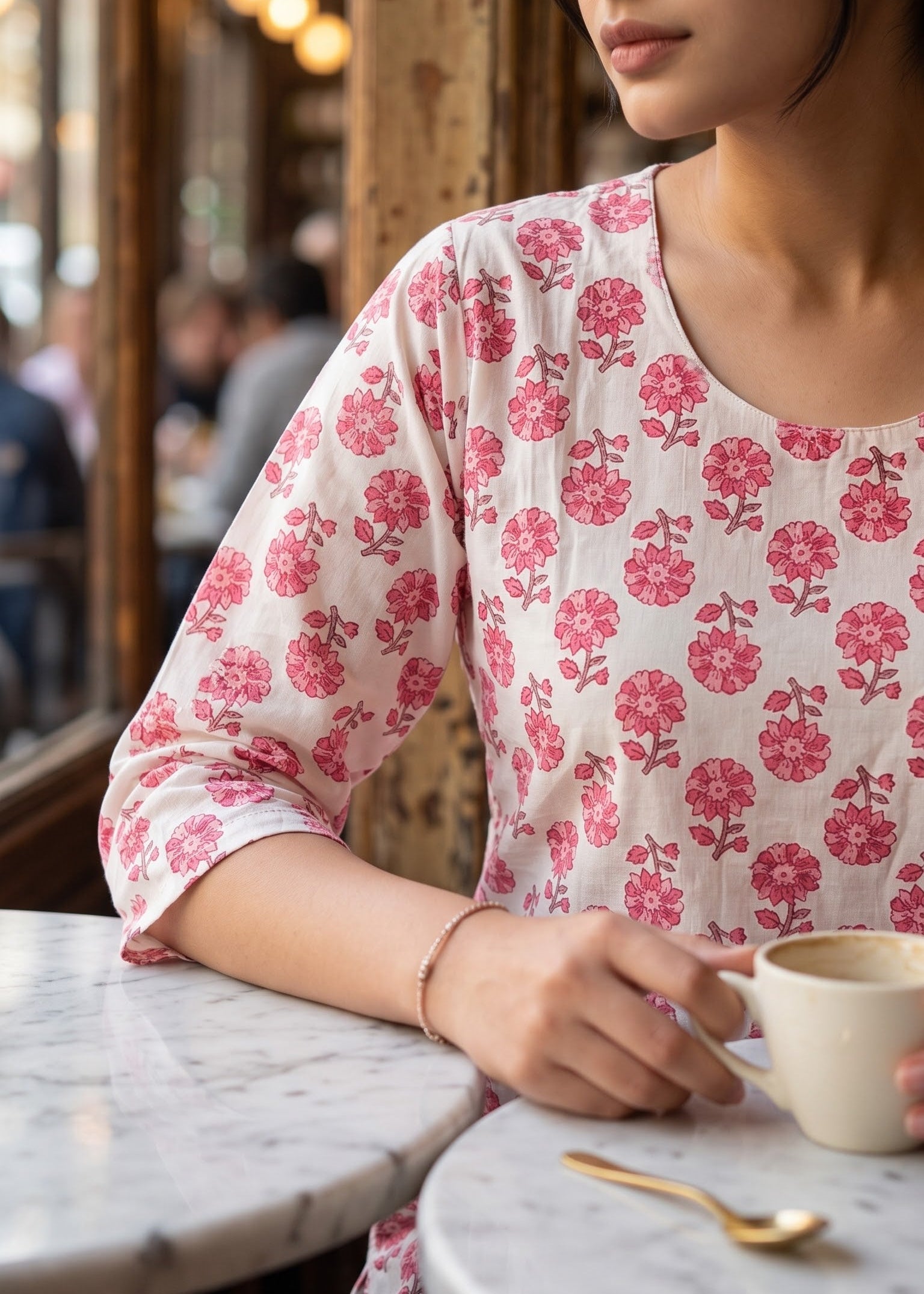 A woman wearing a white short kurta with a pink floral pattern sits at a cafe table, holding a white mug.