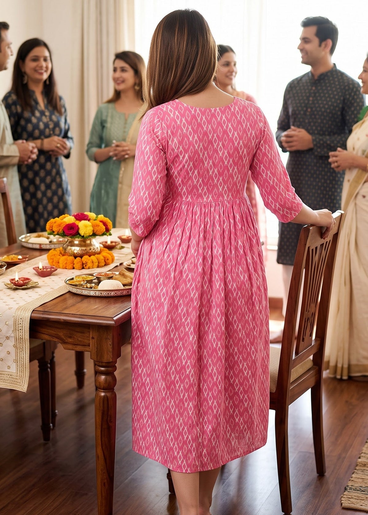 A pregnant woman wearing a pink patterned long-sleeve maternity kurta dress stands with her back to the camera in a dining room.