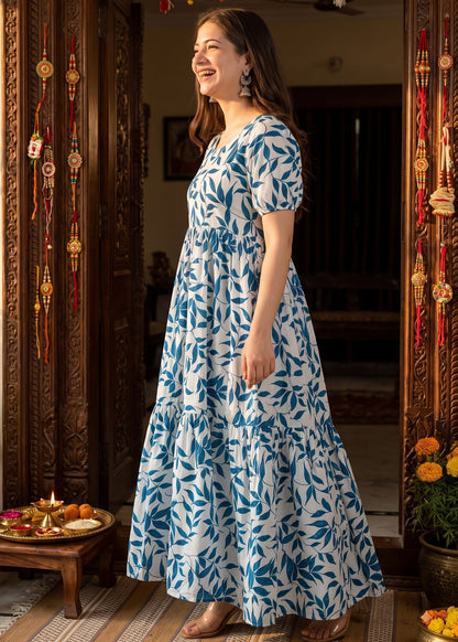 A woman wearing a white short-sleeve maxi dress with a blue leaf pattern stands in a doorway decorated with hanging ornaments and plants.