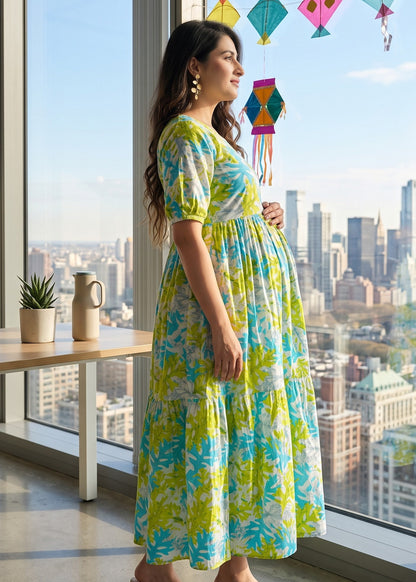A pregnant woman stands by a large window in a city setting, wearing a vibrant yellow and blue floral maxi maternity dress with puff sleeves. 