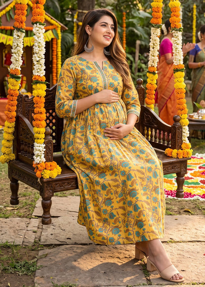 A pregnant woman wearing a yellow and teal floral print maternity kurta dress sits on a wooden swing decorated with marigold garlands in an outdoor garden setting.