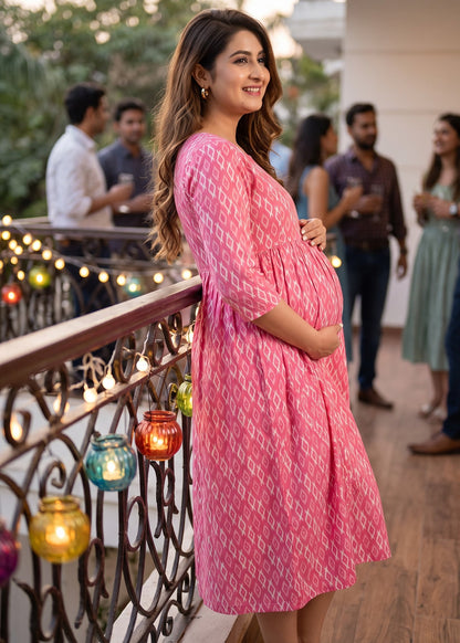 A pregnant woman wearing a pink and white geometric patterned maternity dress stands on a balcony at a social event, holding her baby bump. The dress features a pink and white geometric ikat pattern and three-quarter sleeves.