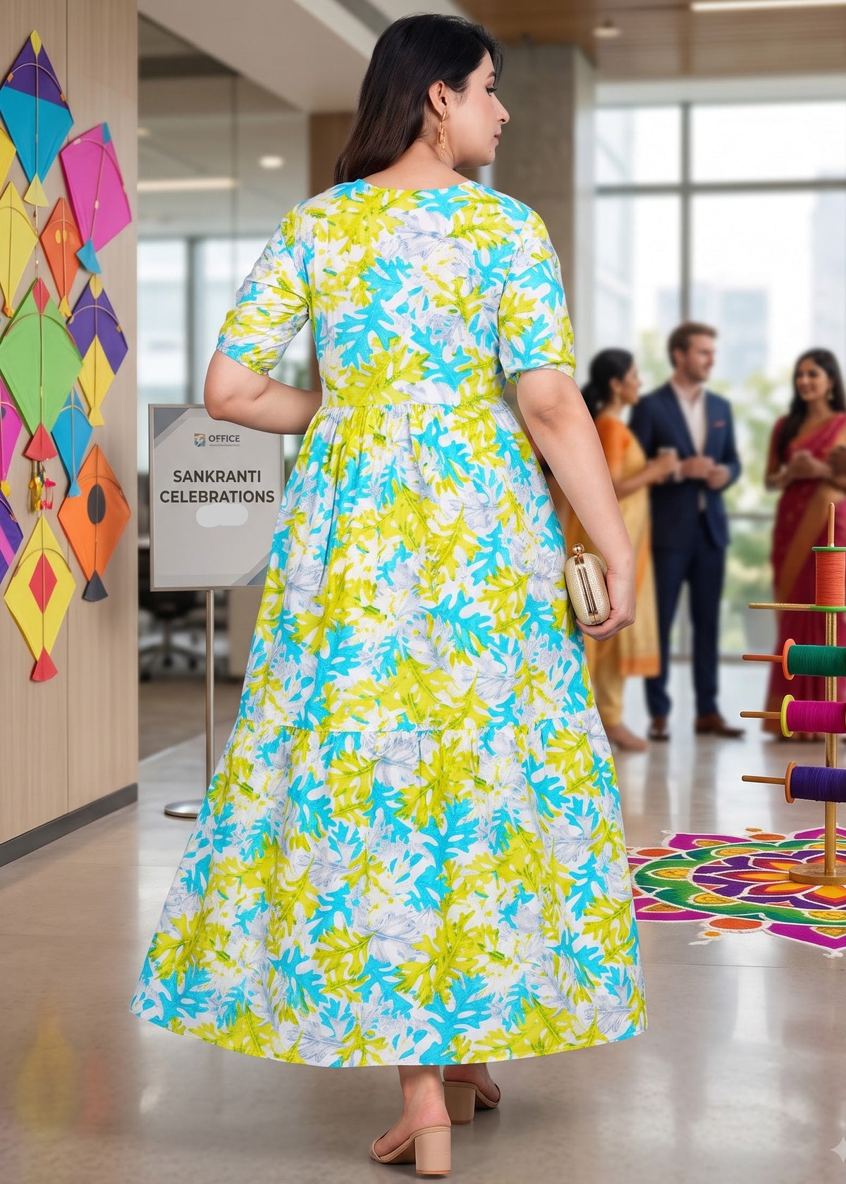 A woman wearing a vibrant yellow, blue, and white floral print maternity midi maxi dress, photographed from behind in a brightly lit office lobby.