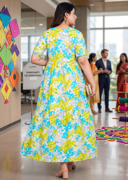 A woman wearing a vibrant yellow, blue, and white floral print maternity midi maxi dress, photographed from behind in a brightly lit office lobby.