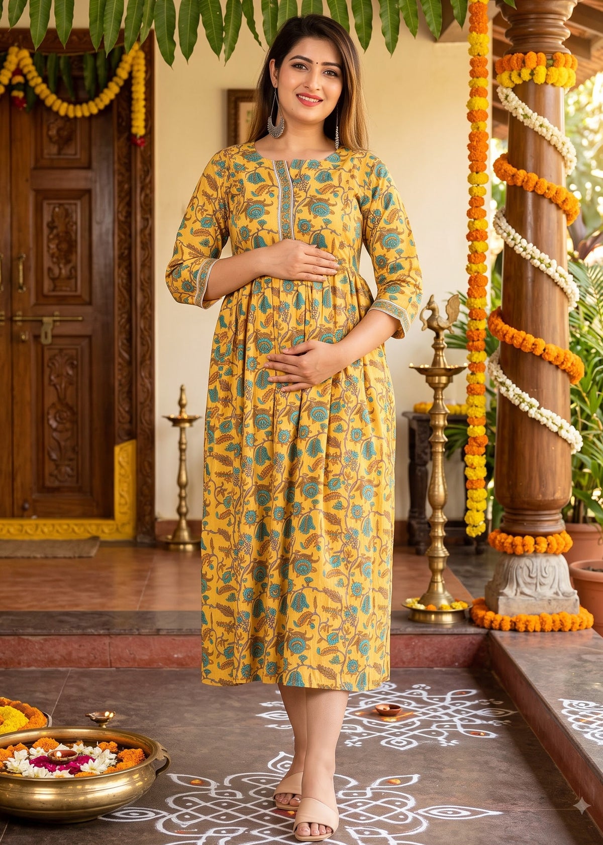 A woman wearing a yellow and teal printed maternity kurta dress stands on a decorated porch with marigold garlands, brass lamps, and floral arrangements.