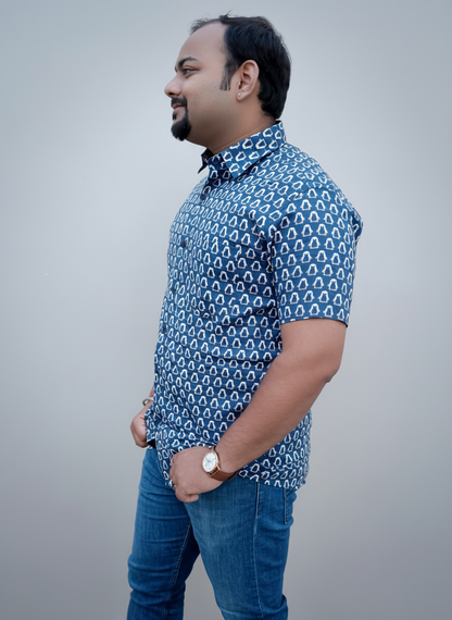 Man wearing a blue patterned shirt and jeans standing against a plain wall.
