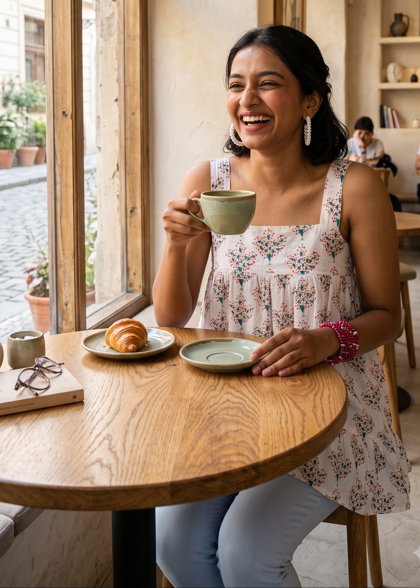 A woman smiling in a cozy cafe setting wearing a sleeveless floral print top and light blue jeans.